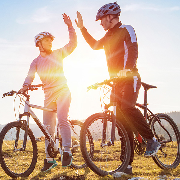 Two people on bicycles, one person waving at the other in a sunlit outdoor setting.