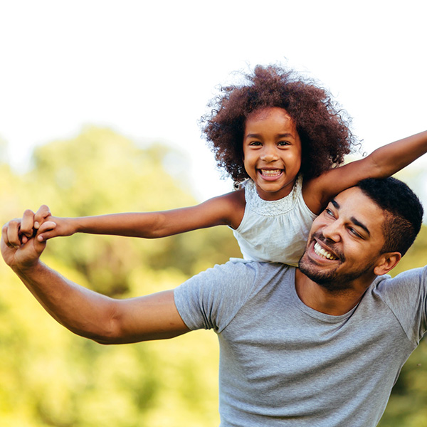 A man and a young girl, both smiling broadly, embracing joyfully in an outdoor setting.