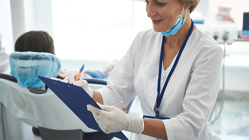 A female dental hygienist in a clinical setting, holding a clipboard and paperwork, wearing gloves and a face mask.