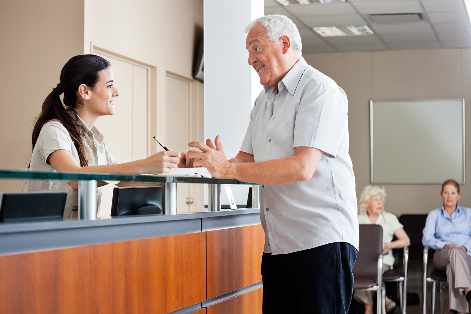 The image depicts an older man and a younger woman in what appears to be a reception area or office, with the man standing behind a counter and the woman seated on the other side.